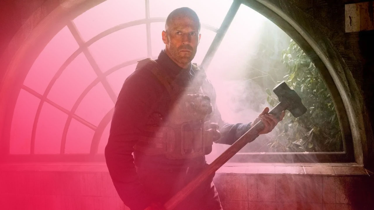 Ein glatzköpfiger Mann mit kurzem Bart und intensivem Blick steht in einer dusteren Umgebung, die von einem großen, halbkreisförmigen Fenster mit Gitterstruktur im Hintergrund beleuchtet wird. Er trägt eine dunkle, taktische Weste über einem dunklen Hemd und hält eine Art Vorschlaghammer über seiner Schulter. Die Beleuchtung ist hell, erzeugt Gegenlicht vom Fenster und lässt die Szene nebelig und dramatisch erscheinen.