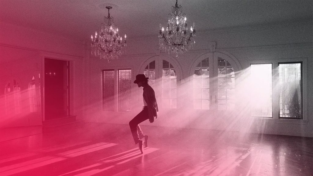 Ein Schwarz-Weiß-Bild zeigt Michael Jackson (oder eine Figur, die Michael Jackson darstellt) in einem leeren Raum, wie er sich im charakteristischen "Anti-Schwerkraft"-Lean nach vorne beugt. Er trägt einen Filzhut, eine Jacke, ein Hemd und Hosen, die eng anliegen und seine schlanke Figur betonen. Seine Beine sind gebeugt, und seine Füße, die wie in speziellen Schuhen stecken, sind fest auf dem Boden verankert, wodurch die Illusion der Unabhängigkeit von der Schwerkraft entsteht. Der Raum ist hell erleuchtet durch Sonnenlicht, das durch mehrere große Fenster auf der rechten Seite fällt und dicke Lichtstrahlen durch den nebligen oder staubigen Raum erzeugt. Zwei große, verzierte Kronleuchter hängen von der Decke, und der Holzboden reflektiert schwach das Licht. Die Szene vermittelt eine dramatische und kunstvolle Atmosphäre.