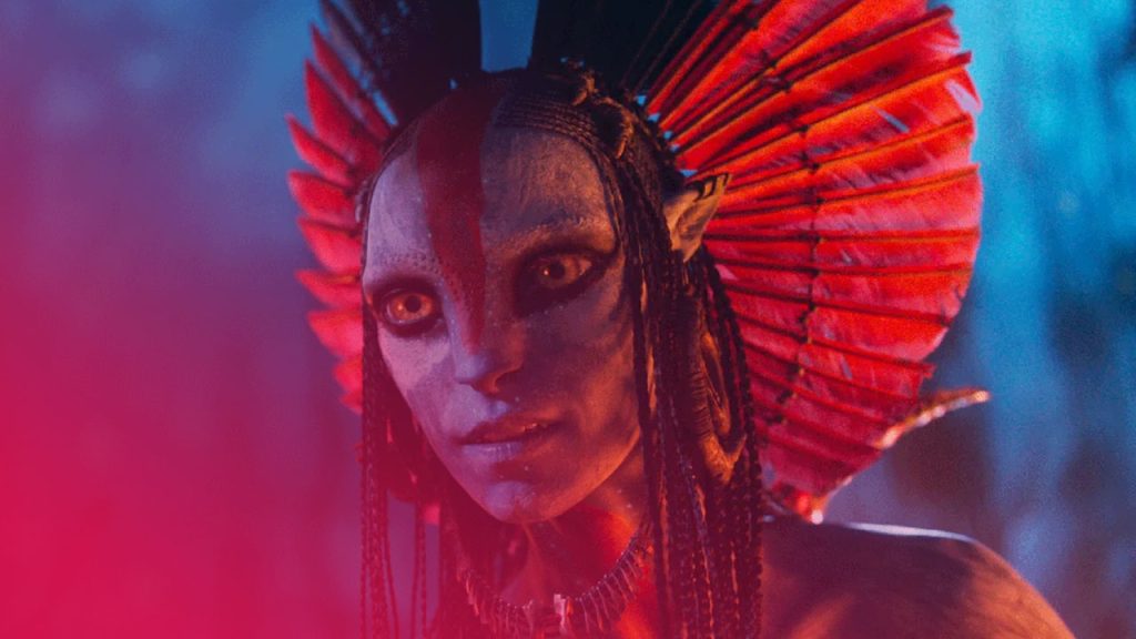 Nahaufnahme des Oberkörpers eines Na'vi, dessen blaue Haut mit roten Stammesmarkierungen auf der Stirn und über die Nase bemalt ist. Die Figur blickt direkt nach vorne mit weit geöffneten, goldenen Augen und trägt einen großen Kopfschmuck aus leuchtend roten Federn oder fächerartigen Elementen, die hinter ihrem Kopf aufgefächert sind. Mehrere schwarze Zöpfe rahmen das Gesicht ein. Der Hintergrund ist ein unscharfes, dunkles Blau, was eine mystische, nächtliche Stimmung erzeugt.