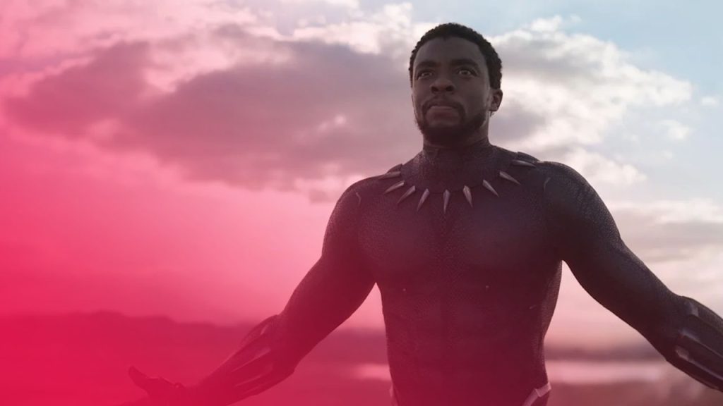 Ein Mann in einem schwarzen Anzug mit silbernen Akzenten steht mit ausgebreiteten Armen vor einem bewölkten Himmel und einer Hügellandschaft.