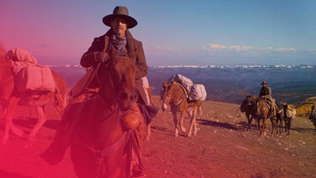 Männer reiten auf Pferden und Maultieren, die mit Packtaschen beladen sind, durch eine hügelige Landschaft unter blauem Himmel.