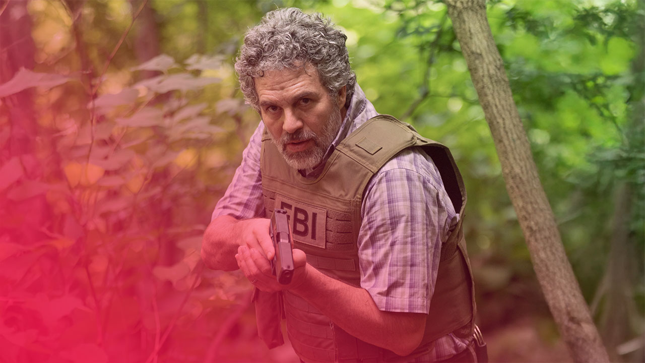 Mann mit grauen Locken und Bart, in Tarnweste mit FBI-Aufschrift, zielt mit Pistole in bewaldeter Umgebung.