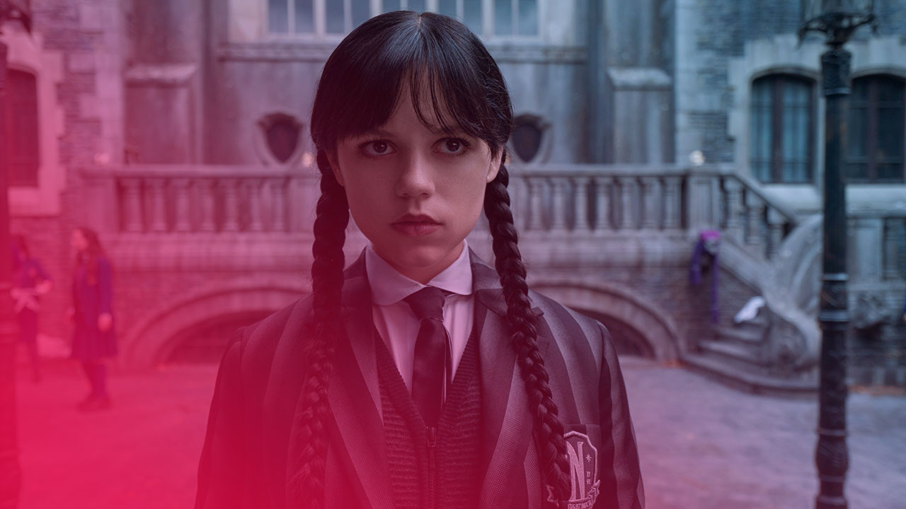 Eine junge Frau mit schwarzen Zöpfen und blasser Haut steht vor einem grauen, gotischen Gebäude. Sie trägt eine gestreifte Schuluniform mit Krawatte und einem Abzeichen. Ihr Blick ist ernst und direkt.