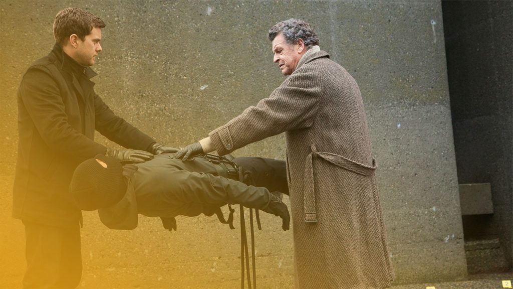 Zwei Männer in Mänteln berühren eine in der Luft schwebende Person, deren Kopf mit einer Sturmhaube bedeckt ist, vor einer Betonwand.