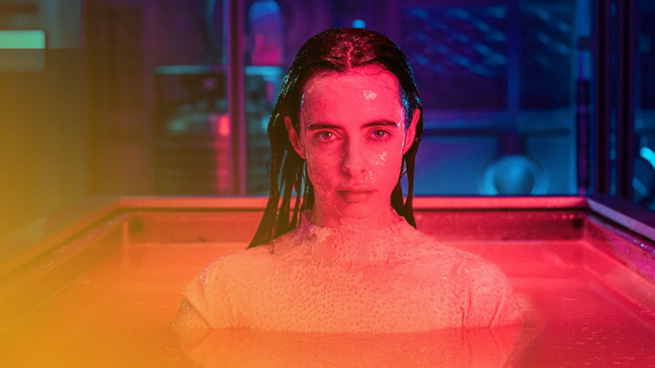 Eine blasse Frau taucht bis zur Brust in rötlicher, fluoreszierender Flüssigkeit, ihr Haar ist nass und nach hinten gekämmt, umgeben von einem dunklen, futuristisch beleuchteten Raum in Neonrosa und Blau.