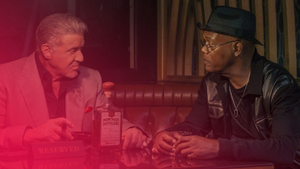 Zwei Männer sitzen an einem Tisch mit Whiskeyflasche und Gläsern in einer Bar. Einer trägt einen Hut. Ein Schild "Reserved" steht auf dem glänzenden Tisch.