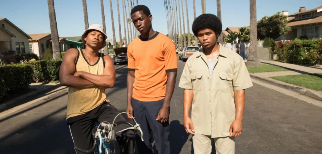 Drei junge Männer stehen auf einer sonnigen, von Palmen gesäumten Straße; der mittlere trägt ein orangefarbenes T-Shirt.