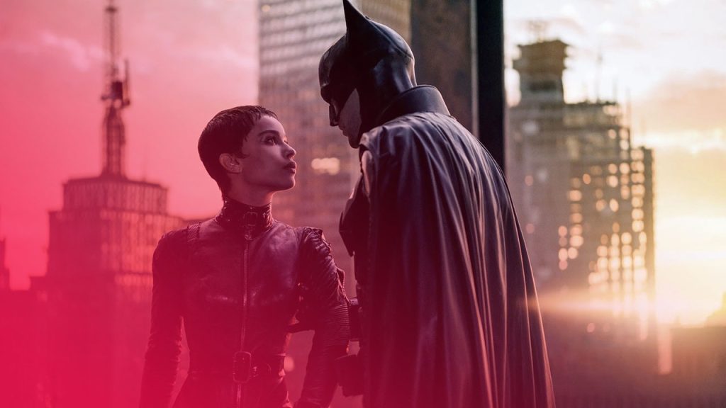 Eine Frau in schwarzem Lederanzug blickt zu einem Mann im Batman-Kostüm auf. Sie stehen auf einem Dach vor einer Stadtkulisse bei Dämmerung mit orange-goldenem Himmel.