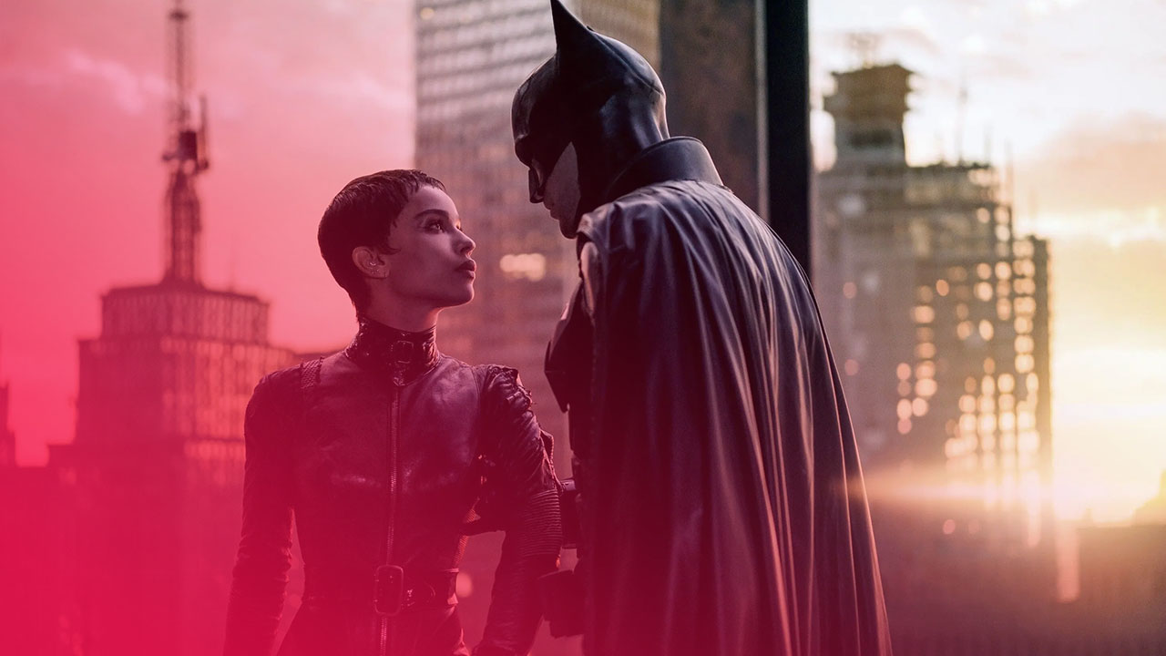 Eine Frau in schwarzem Lederanzug blickt zu einem Mann im Batman-Kostüm auf. Sie stehen auf einem Dach vor einer Stadtkulisse bei Dämmerung mit orange-goldenem Himmel.