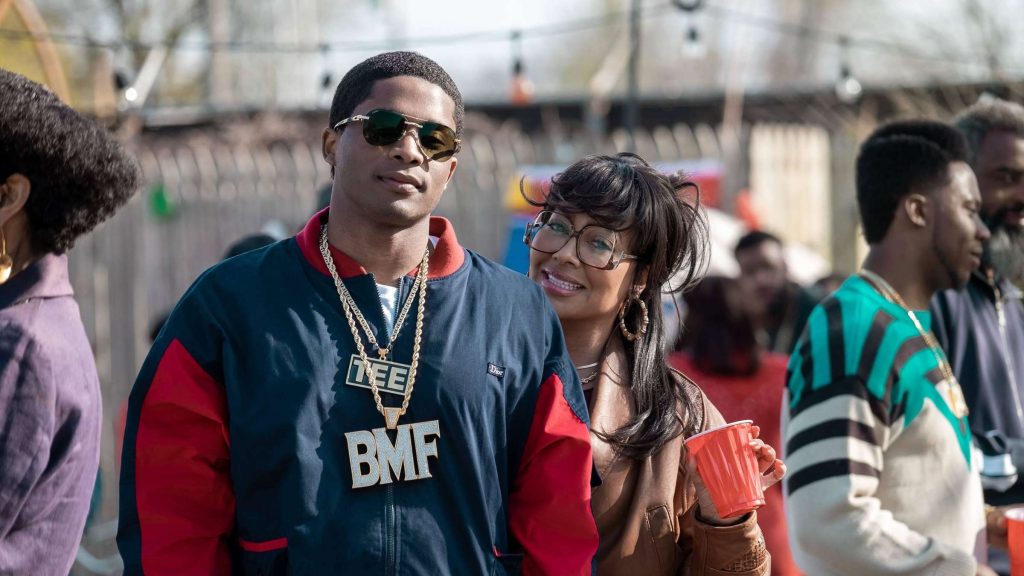 Nahaufnahme von zwei Darstellern aus der Serie BMF, die im Freien stehen. Der Mann in der marineblauen und roten Jacke trägt große goldene „TEE“- und „BMF“-Ketten und eine Sonnenbrille. Die Frau lehnt lächelnd an seiner Schulter, trägt eine braune Lederjacke, eine Brille und hält einen roten Becher.