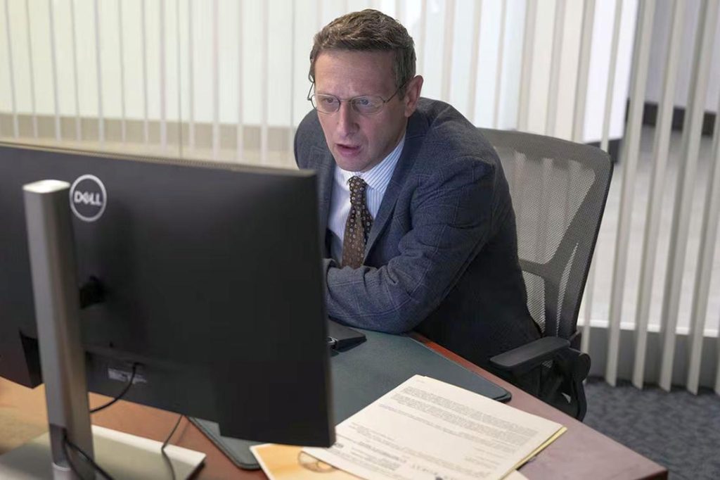 Ein Mann in Anzug und Brille sitzt an einem Schreibtisch vor einem Dell-Monitor, schaut überrascht auf den Bildschirm. Büroumgebung.