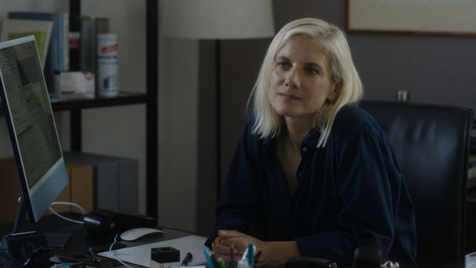 Frau mit blondem Haar sitzt an einem dunklen Schreibtisch mit Computer und Büromaterial. Sie trägt ein blaues Hemd und blickt nach links. Im Hintergrund ein Regal und eine Lampe.