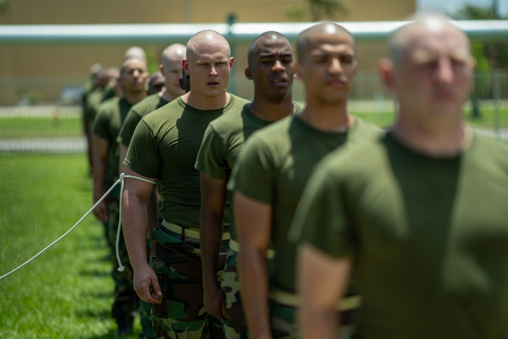 Männer mit rasierten Köpfen in grünen T-Shirts und Tarnhosen stehen im Freien in einer Reihe hintereinander stramm.