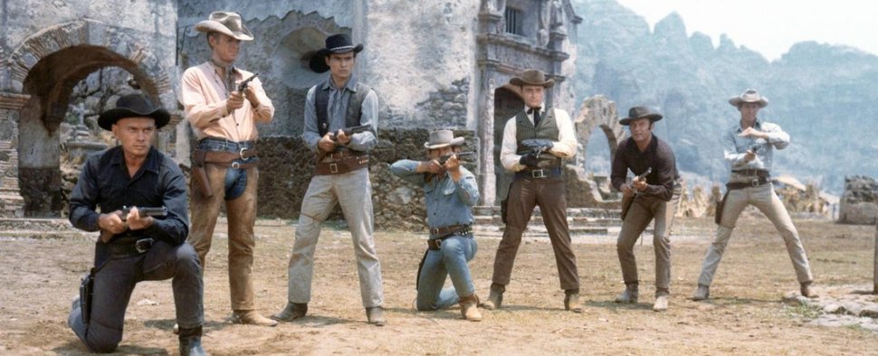 Sieben Cowboys posieren mit gezogenen Revolvern und Gewehren vor einer rustikalen Steinkulisse im Wilden Westen.