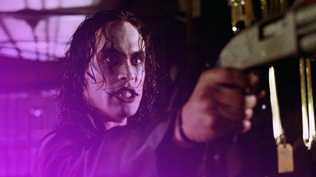 Mann mit bleichem Gesicht, dunkler Schminke und langen, dunklen, nassen Haaren zielt mit einer Pistole in dunkler Umgebung.