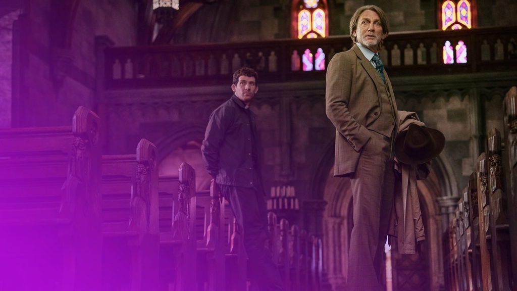 Zwei Männer in einer dunklen, gotischen Kirche mit Buntglasfenstern. Rechts ein Mann im braunen Anzug mit Hut, links ein Mann in dunkler Jacke.