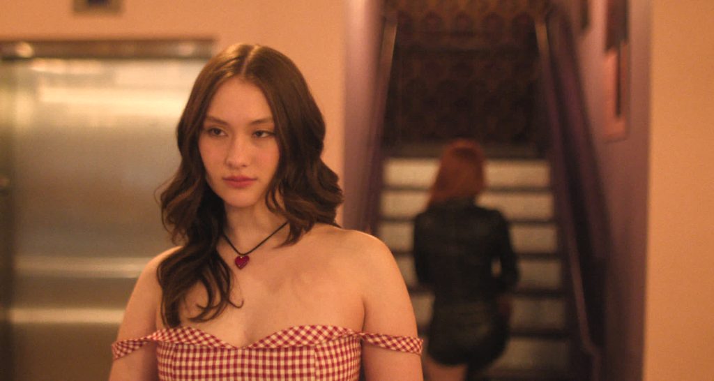 Frau in rot-weiß kariertem Kleid und Herzanhänger blickt ernst. Im Hintergrund Treppe und unscharfe Person in Schwarz.