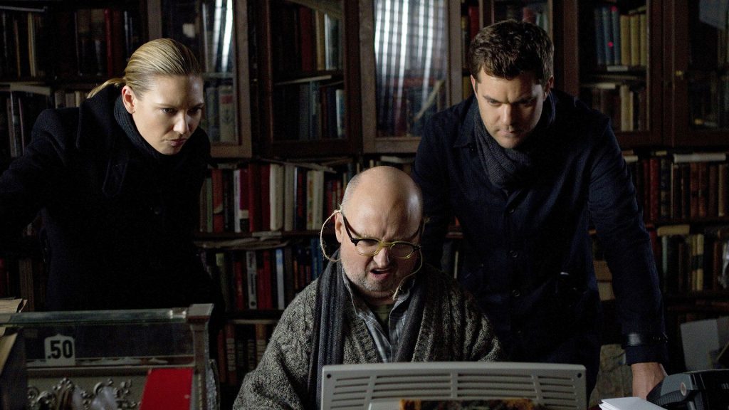 In einer Bibliothek stehen drei Personen vor einem alten Computer. Eine Frau und ein Mann blicken auf den Bildschirm, während ein Mann mit Brille tippt.