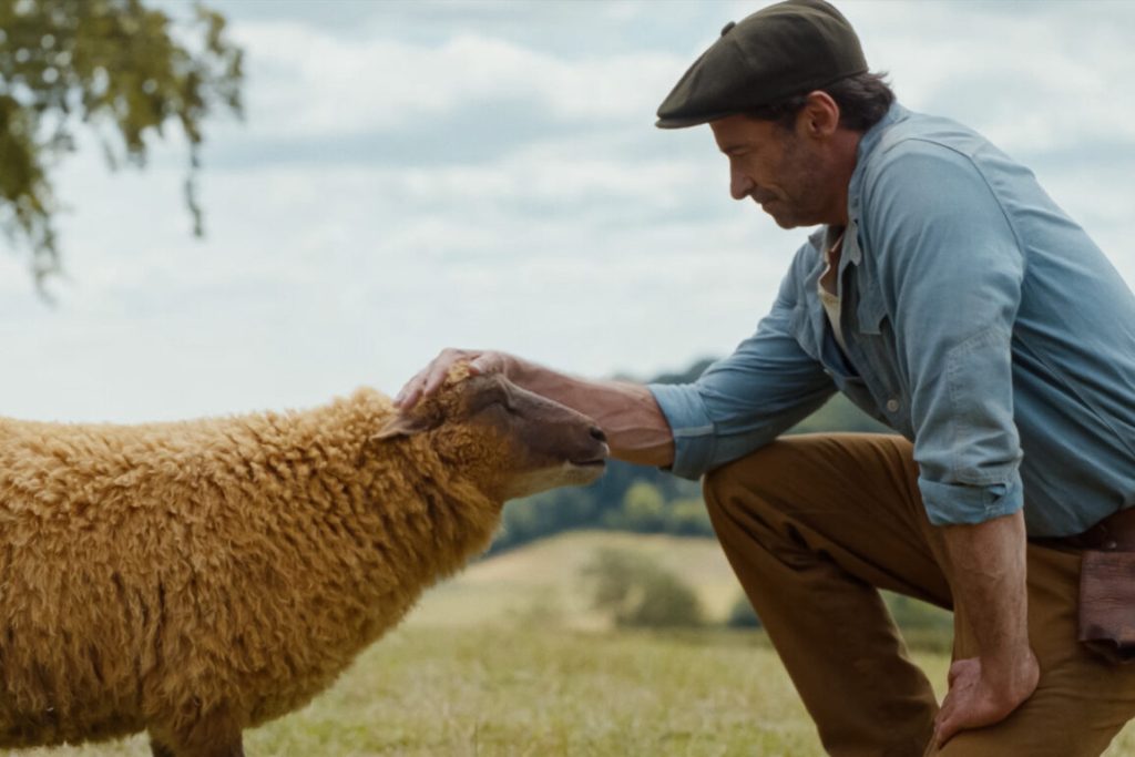 Ein Mann in Arbeitskleidung und Schiebermütze kniet auf einer Wiese und streichelt sanft den Kopf eines hellbraunen Schafes.