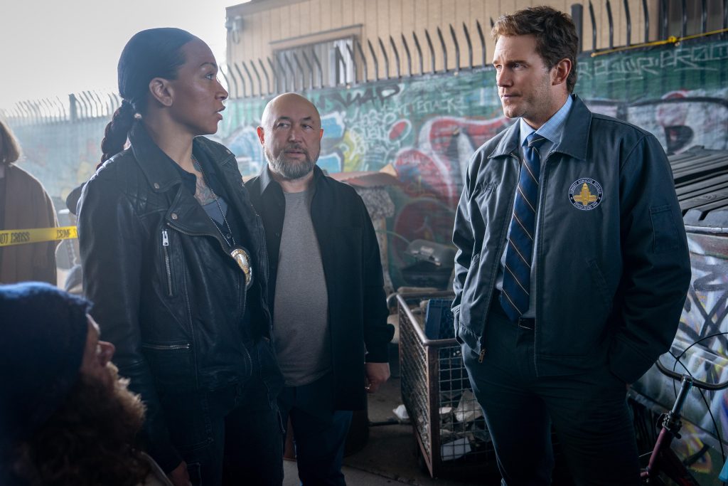 Drei Personen, darunter zwei Beamte mit Abzeichen, sprechen ernsthaft in einer graffitibedeckten Stadtkulisse. Gelbes Absperrband, Müll, Fahrrad sichtbar.