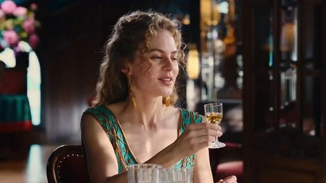 Frau mit blonden Locken und Ohrringen hält ein Glas mit bernsteinfarbener Flüssigkeit. Sie trägt ein grünes, verziertes Oberteil. Hintergrund: dunkles Interieur, Blumen.