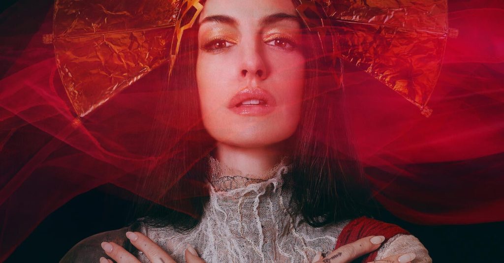 Frau mit goldenem Kopfschmuck und goldenem Lidschatten vor rotem Schleier. Hände gekreuzt, Tattoos sichtbar. Blasses Kleid mit Spitze.