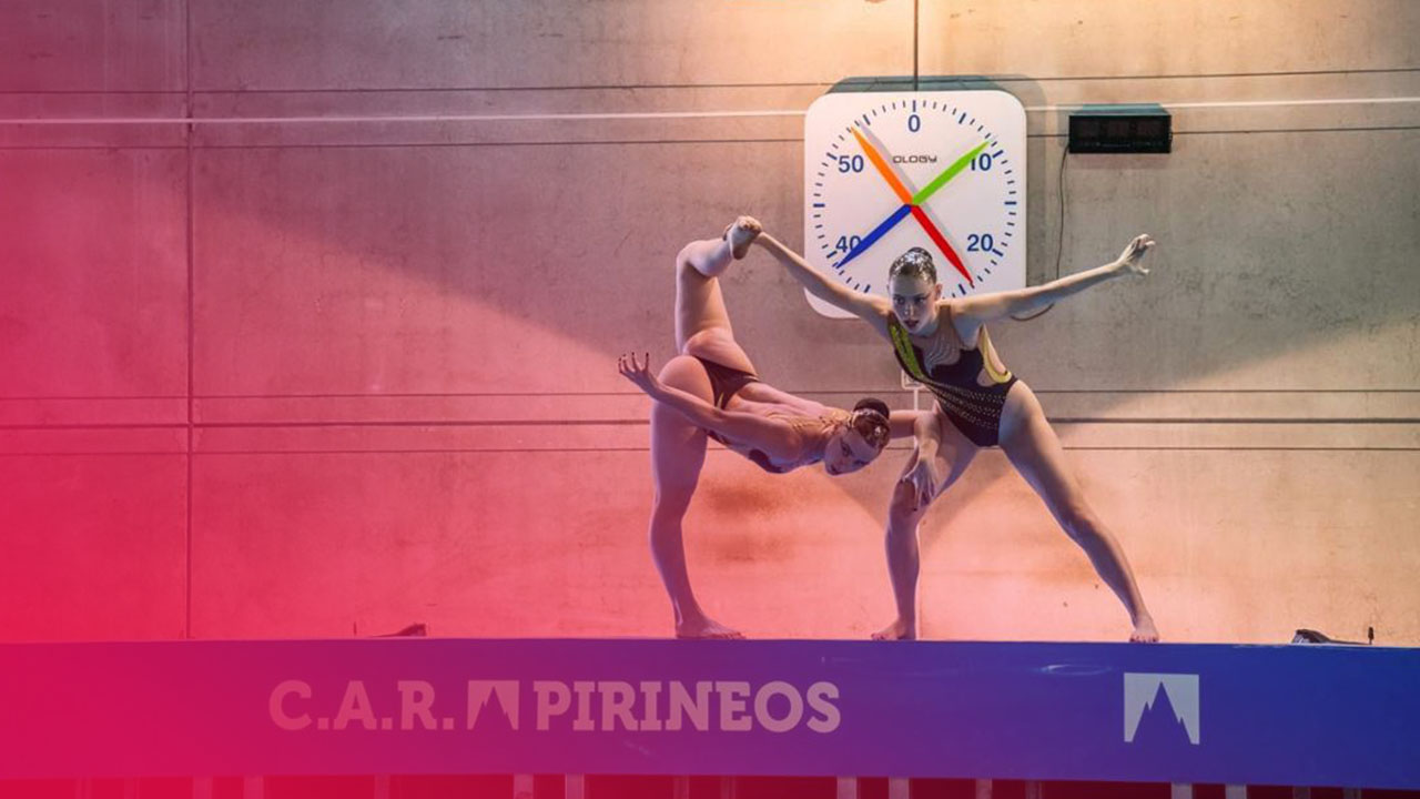 Zwei Synchronschwimmerinnen posieren in akrobatischen Übungen am Beckenrand des C.A.R. Pirineos vor einer großen Uhr.