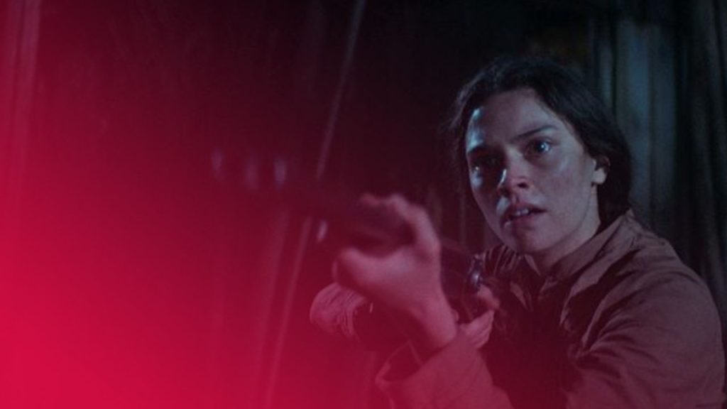 Eine Filmszene bei Nacht oder in einer dunklen Umgebung. Eine junge Frau mit dunklen Haaren und einem beigefarbenen oder hellbraunen Hemd hält ein Gewehr auf Schulterhöhe, zielt und blickt mit einem entschlossenen, leicht besorgten Ausdruck in die Ferne. Ihr Gesicht ist rechts im Bild hell beleuchtet, während der Hintergrund links dunkel und unscharf ist, was eine angespannte, dramatische Stimmung erzeugt.