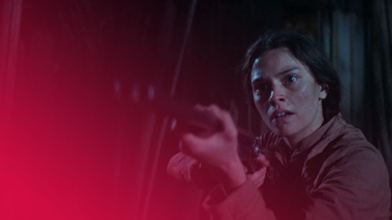 Eine Filmszene bei Nacht oder in einer dunklen Umgebung. Eine junge Frau mit dunklen Haaren und einem beigefarbenen oder hellbraunen Hemd hält ein Gewehr auf Schulterhöhe, zielt und blickt mit einem entschlossenen, leicht besorgten Ausdruck in die Ferne. Ihr Gesicht ist rechts im Bild hell beleuchtet, während der Hintergrund links dunkel und unscharf ist, was eine angespannte, dramatische Stimmung erzeugt.