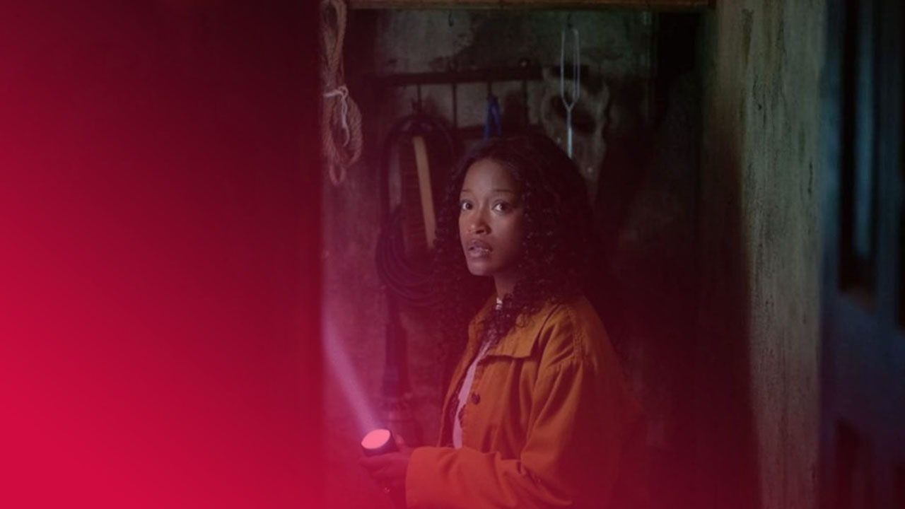 Eine junge Frau mit lockigem Haar und gelber Jacke steht in einem dunklen Raum und leuchtet mit einer Taschenlampe. Ihr Blick ist ängstlich. An der Wand hängen Werkzeuge und ein Schädel.