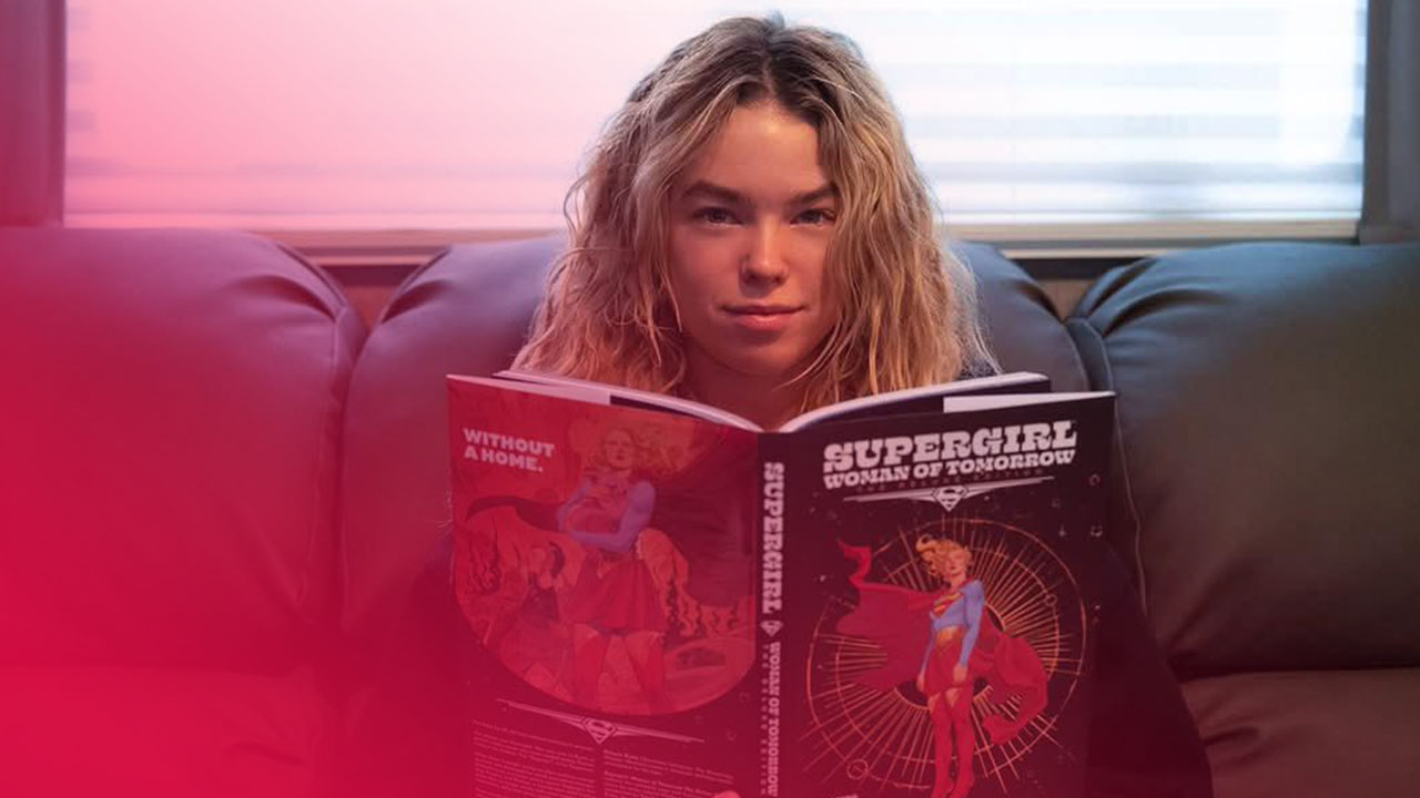 Blonde Frau auf dunklem Sofa hält geöffneten "Supergirl: Woman of Tomorrow" Comic mit Titelheldin auf beiden Seiten, blickt in die Kamera.