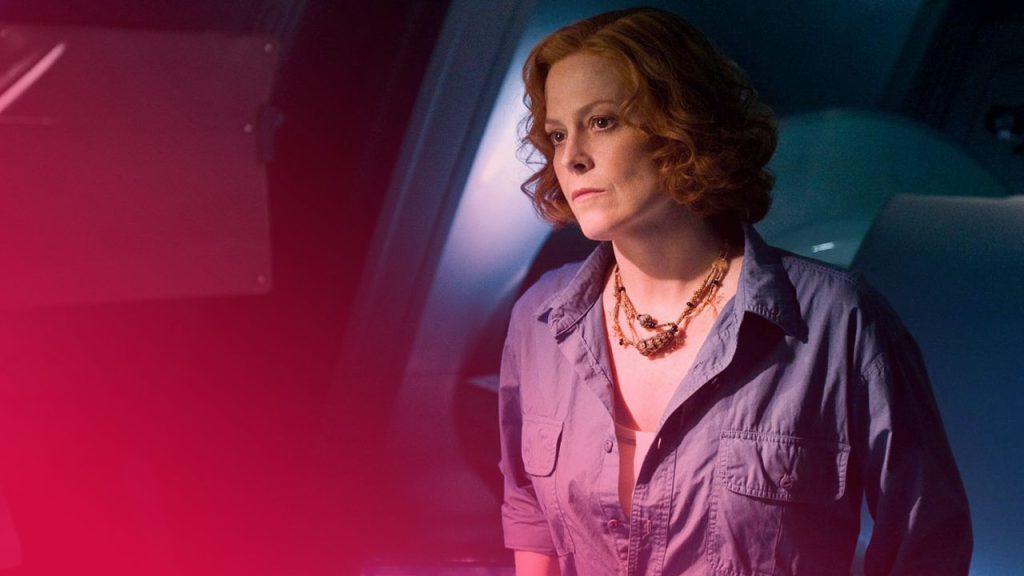 Nahaufnahme einer nachdenklichen Frau mit rötlichem Haar, blauer Bluse und mehrsträngiger Halskette in einem dunkelblauen Raum.