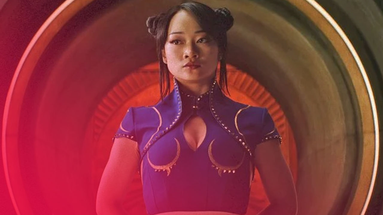 Asiatische Frau mit zwei Haarknoten, ernstem Blick, in blauem Oberteil mit Goldakzenten vor dunklem, ringförmigem Tunnel mit orangefarbenem Licht.