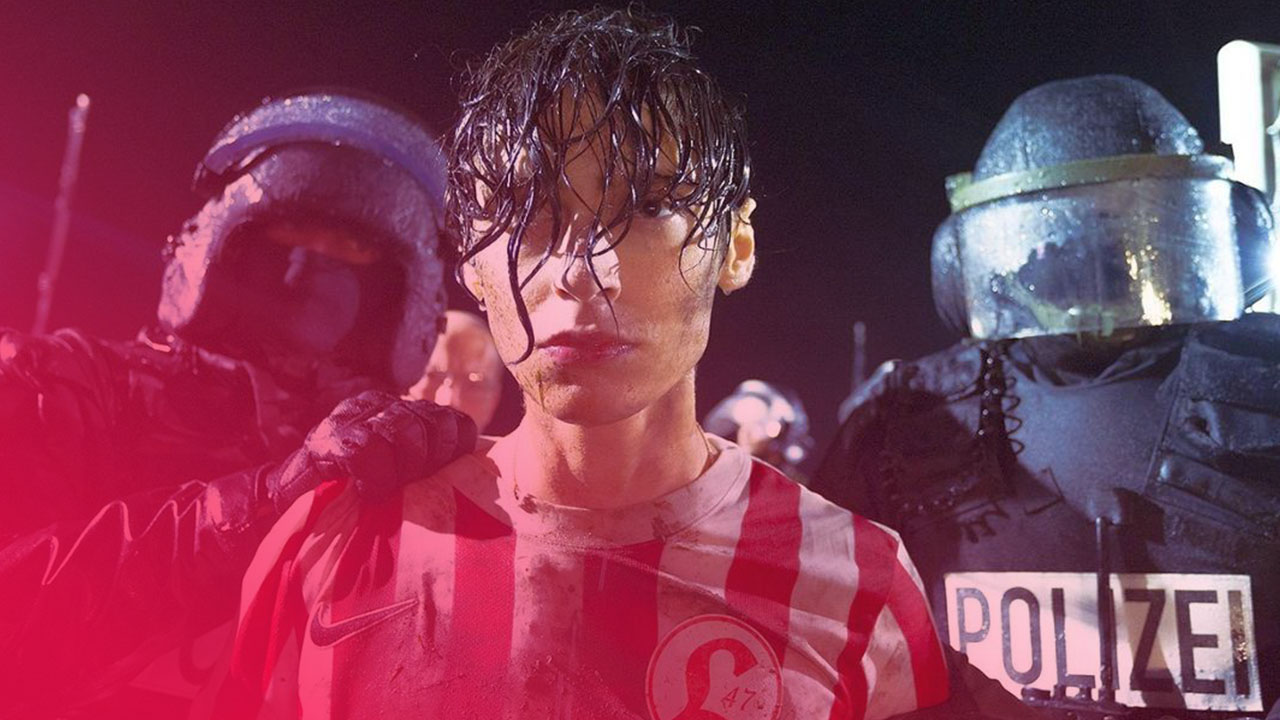 Ein nasser Jugendlicher in rot-weißem Trikot, Gesicht schmutzig, wird von zwei Polizisten in voller Montur festgehalten. Dunkle, angespannte Nachtszene.