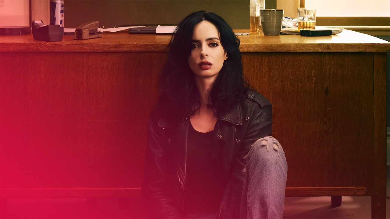 Frau mit dunklen Haaren, Lederjacke und zerrissenen Jeans sitzt vor einem Schreibtisch mit Akten und Gläsern.