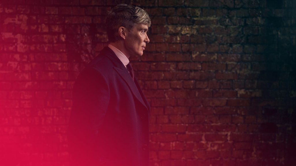 Ein Mann im dunklen Mantel, Hemd und Krawatte steht vor einer roten Backsteinmauer. Er blickt nach rechts. Das Bild hat dunkle Töne.