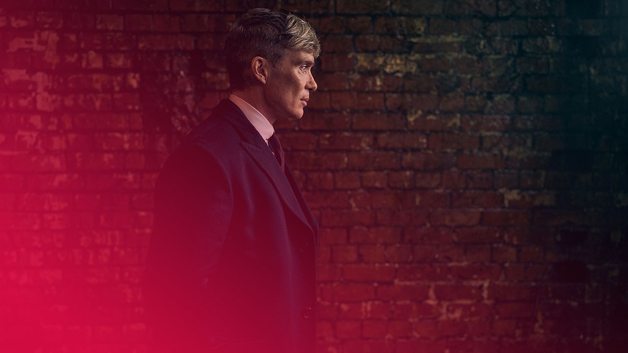 Ein Mann im dunklen Mantel, Hemd und Krawatte steht vor einer roten Backsteinmauer. Er blickt nach rechts. Das Bild hat dunkle Töne.