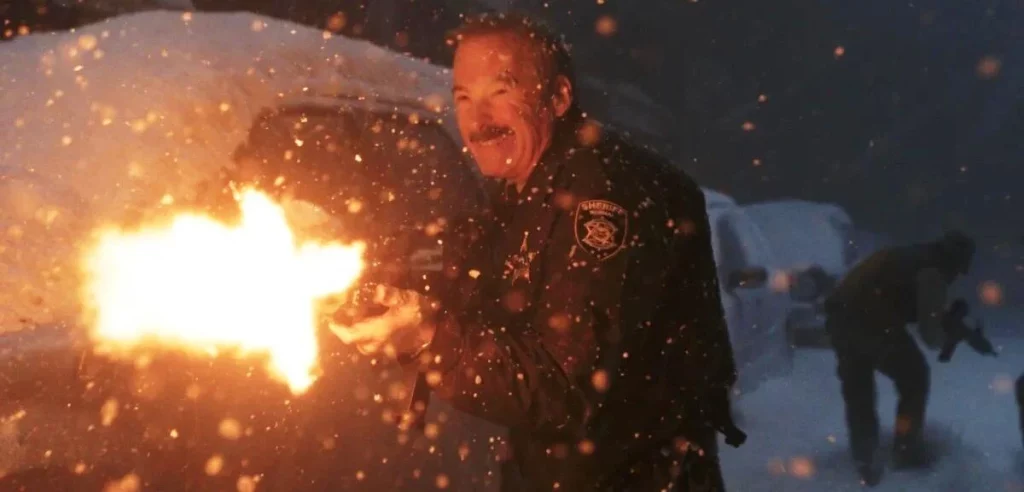 Ein Sheriff mit angespannter Miene feuert eine Waffe ab, erzeugt helles Mündungsfeuer. Funken fliegen im dunklen, verschneiten Umfeld. Eine Person kauert hinten.