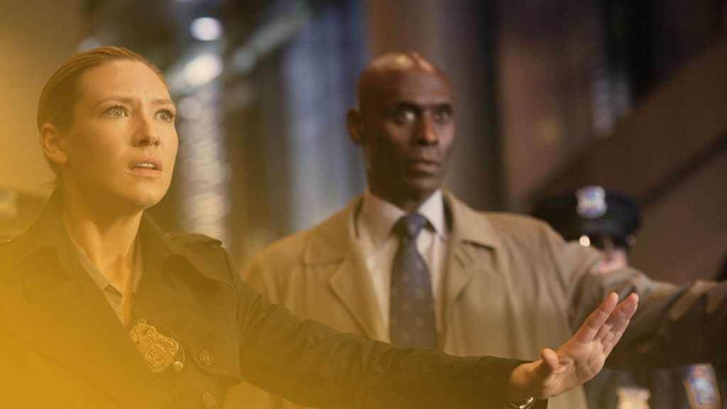 Polizistin in dunkler Jacke, mit Abzeichen, blickt besorgt nach links und hält Stopp-Geste. Mann mit Mantel und Krawatte im Hintergrund. Weitere Polizisten sichtbar.