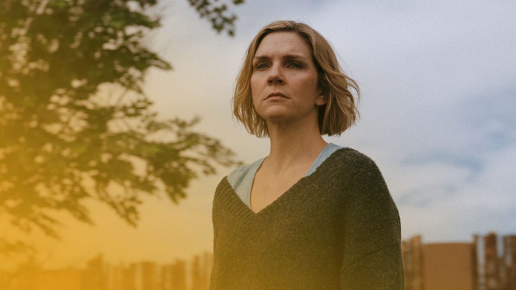 Eine blonde Frau blickt ernst vor einem Baum und Holzzaun unter einem weitläufigen, bewölkten Himmel.