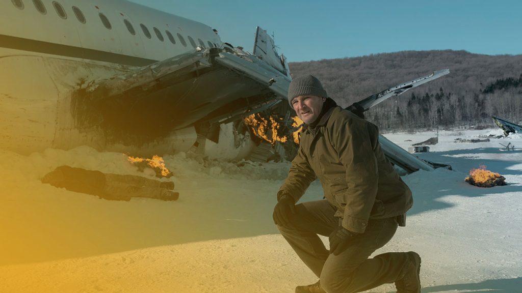 Ein Mann in olivefarbener Jacke und grauer Mütze kniet im Schnee vor einem brennenden Flugzeugwrack. Dahinter kahle Bäume und blauer Himmel.