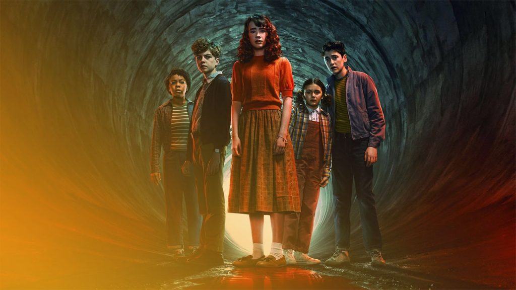 Fünf verängstigte Jugendliche stehen in einem dunklen Tunnel vor einem hellen Ausgang; rotes Licht spiegelt sich am Boden.