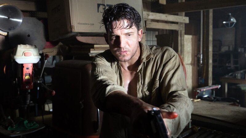 Ein Mann mit dunklem Haar in einem schmutzigen Hemd hält eine Waffe in einer Werkstatt. Er hat einen Kratzer im Gesicht. Werkzeuge und Kisten sind im Hintergrund zu sehen.