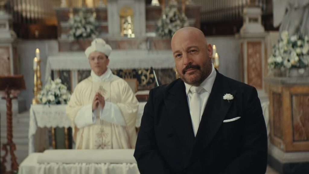 Ein glatzköpfiger, bärtiger Mann im Anzug lächelt in einer Kirche. Im unscharfen Hintergrund steht ein Priester am Altar.