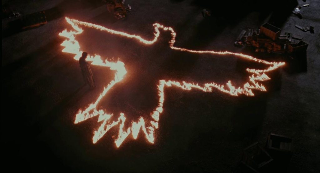 Dunkle Aufnahme von oben: Eine Person steht neben einem Vogelschattenriss aus Feuer auf einer dunklen Oberfläche; Kisten und Müll liegen herum.