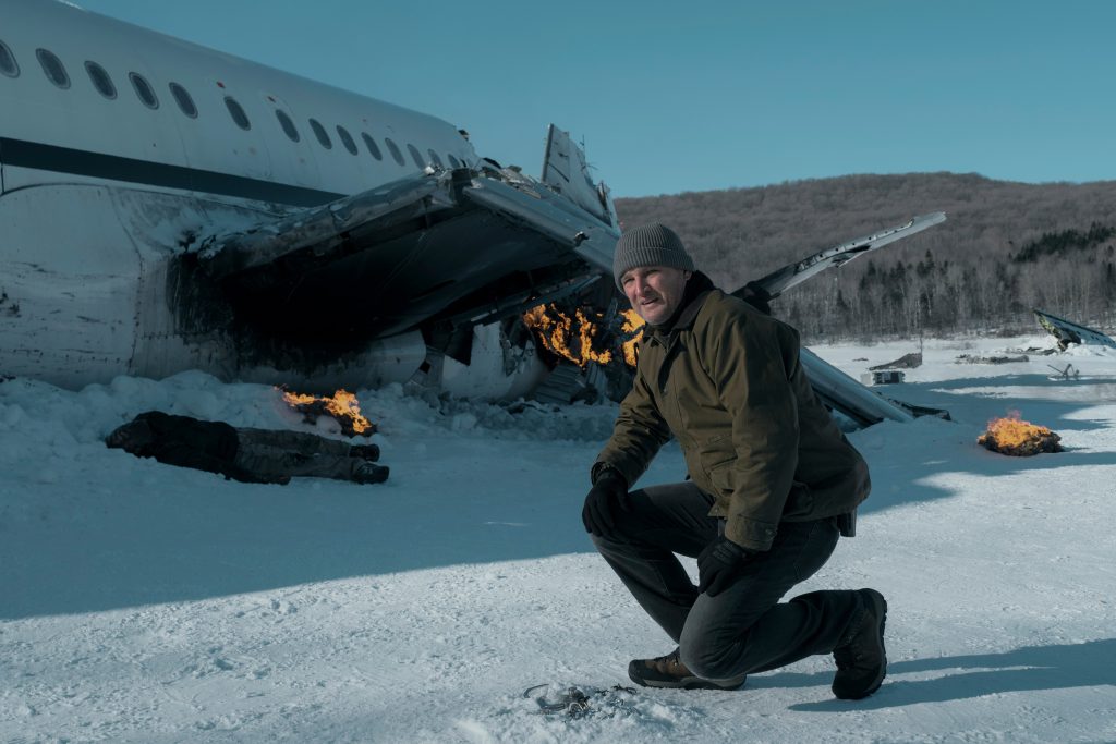 Ein Mann in olivefarbener Jacke und grauer Mütze kniet im Schnee vor einem brennenden Flugzeugwrack. Dahinter kahle Bäume und blauer Himmel.