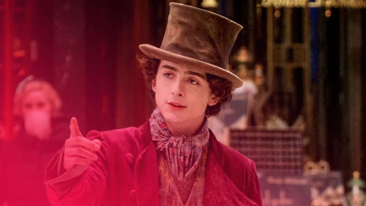 Timothée Chalamet als Willy Wonka im lila Gehrock und Zylinder zeigt mit dem Finger nach vorn in einer Stadtkulisse.