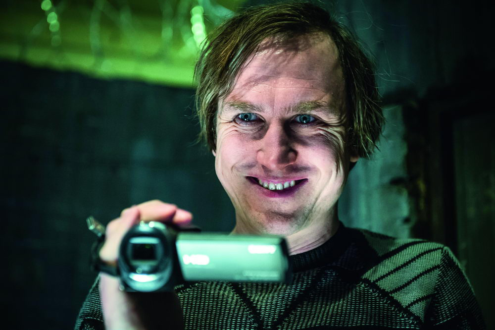 Ein Mann mit manischem Grinsen hält eine Videokamera in einem düsteren, grünlich beleuchteten Raum vor sein Gesicht.