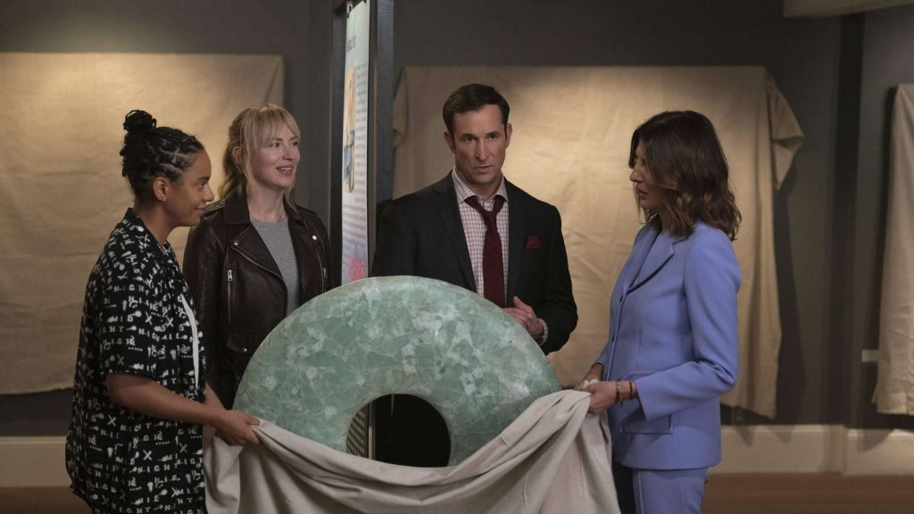 Vier Personen enthüllen konzentriert in einer Ausstellung ein großes, hellgrünes, gemustertes Ringobjekt unter einem beigen Tuch.