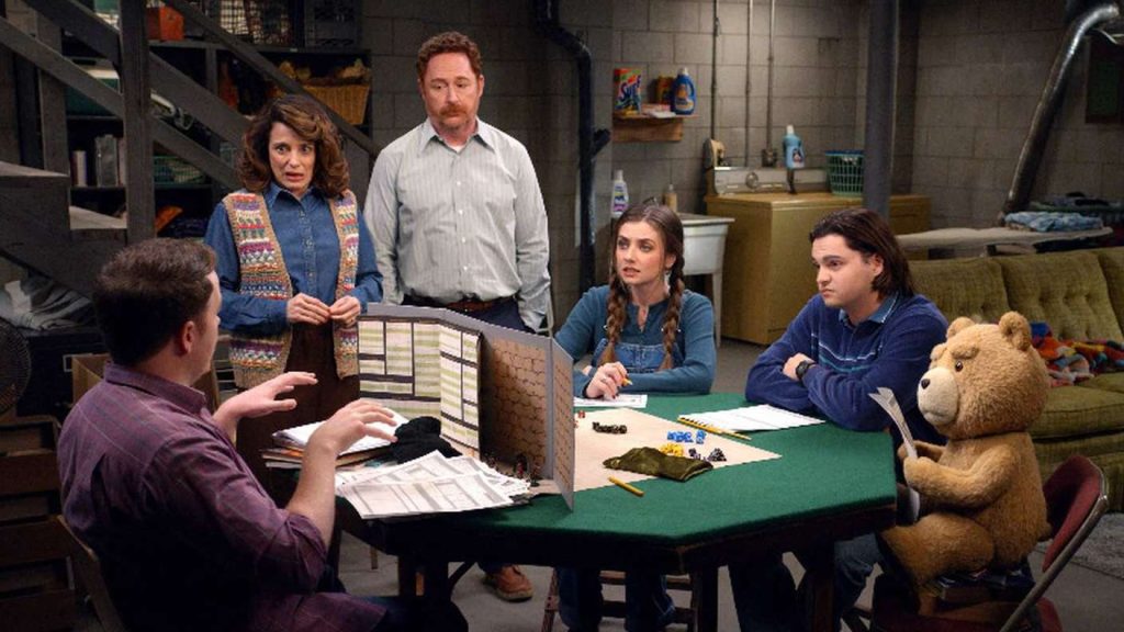 Fünf Personen, ein Teddybär, spielen RPG an grünem Tisch im Keller. Zwei Erwachsene schauen ernst zu. Würfel, Spielleiter-Schild.